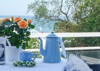 Tisch auf Balkon mit Blick zur Ostsee, auf dem Tisch meerfarbenes Kaffeegeschirr und aprikotfarbener Rosenstrauß
